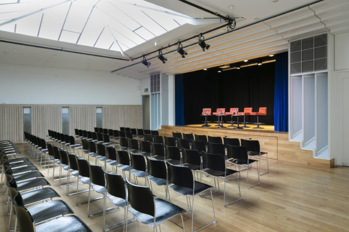 Konzertbestuhlung im Saal. Auf der Bühne stehen fünf rote Barhocker.