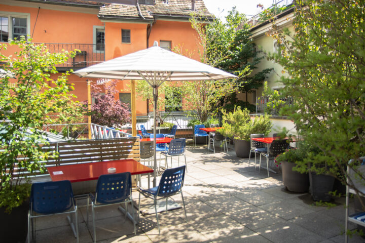 Der Innenhof ist ausgestattet mit Tischen und Stühlen. Es gibt Sonnenschirme und Bäume, die Schatten spenden.