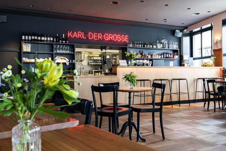 Innenansicht unseres Bistros. Im Bild ist die Bar mit Barhockern. Eine Auswahl aus dem Getränkeangebot ist sichtbar und an der Wand steht in roten Neonbuchstaben "Karl der Grosse". Auf den Bistrotischen stehen Blumen in Glasvasen.