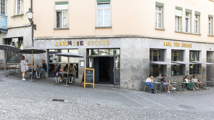 Aussenansicht des Debattierhauses Karl der Grosse. Es gibt genügend Sitzmöglichkeiten und Sonnenschirme, um draussen etwas zu trinken oder zu essen.