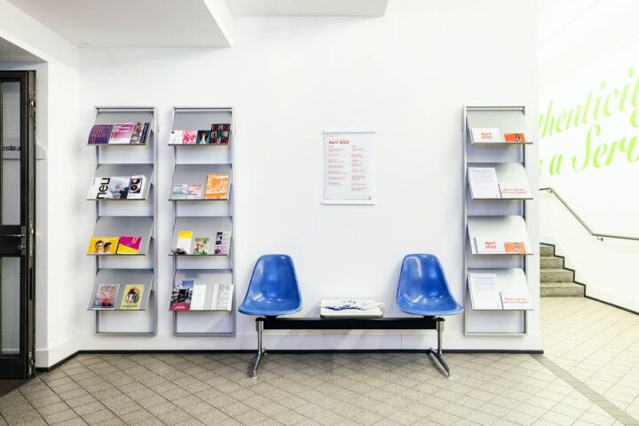 Im Eingangsbereich hängt ein Plakat mit dem Monatsprogramm an der Wand. Drei Flyerständer machen auf Veranstaltungen aufmerksam. Zwei blaue Plastikschalenstühle bieten Sitzmöglichkeiten.