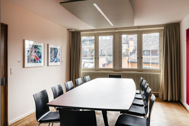 Das Hofblickzimmer ist ausgestattet mit Fischgrätenparkett und einem langen Tisch mit Stühlen rundherum. Ein grosses Fenster an der hinteren Wand bietet viel Licht.