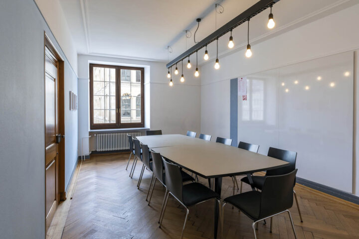 Das Turmblickzimmer hat ein grosses Fenster und Fischgrätenparkett. An der Wand gibt es ein Whiteboard. Mittig steht ein langer Tisch mit Stühlen rundherum und an der Decke hängt eine Schiene, aus der Glühbirnen an Kabeln baumeln.