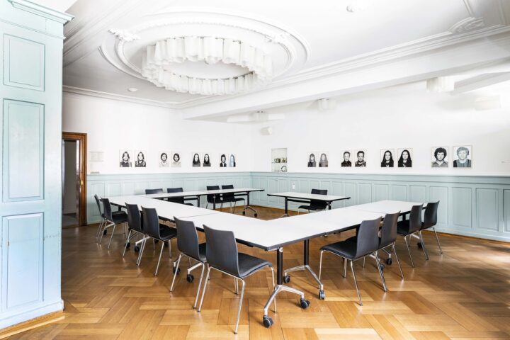 Das Erkerzimmers hat Fischgrätenparkett und hellblaue Wandtäfelungen. Die Tische sind in einer U-Form angeordnet. An den Wänden hängen Fotos der Dauerausstellung "It's a… Women's Women's Women's World!".