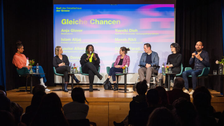 Podiumsdiskussion auf der Bühne im Saal zum Thema "Gleiche Chancen". 7 Personen aus unterschiedlichen Branchen sprechen miteinander. Ein Gebärdensprachdolmetscher übersetzt das Gespräch.