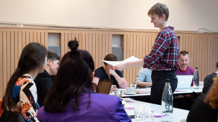 Einblick in einen Workshop. Personen die am Laptop arbeiten. Kaffeetassen und Wassergläser stehen auf dem Tisch. Eine Person teilt Blätter aus.