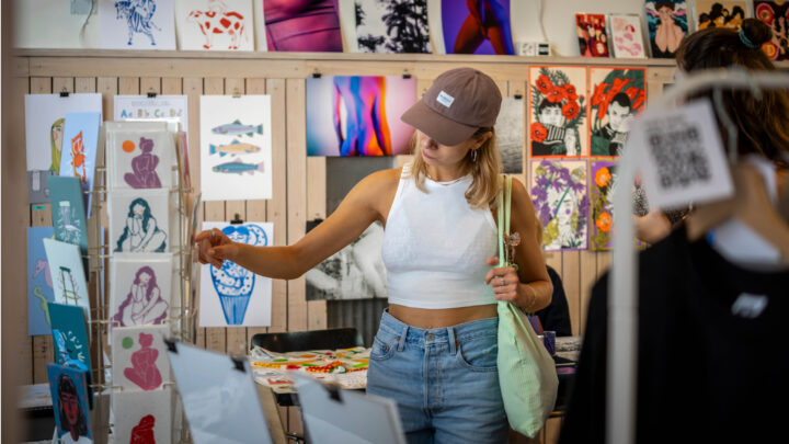 Eine Momentaufnahme des Kunstmarkts. Postkarten und Plakate mit verschiedenen Illustrationen hängen an den Wänden. Eine Frau sieht sich den Postkartenständer an.