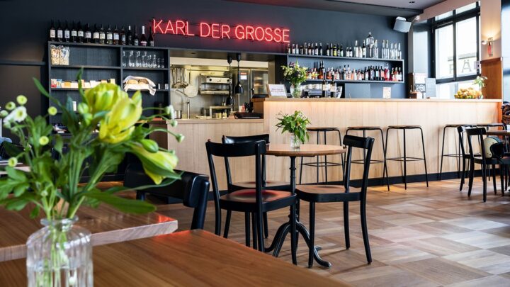 Innenansicht unseres Bistros. Im Bild ist die Bar mit Barhockern, wo man eine Auswahl aus dem Getränkeangebot sieht und an der Wand steht in roten Neonbuchstaben "Karl der Grosse". Auf den Bistrotischen stehen Blumen in Glasvasen.